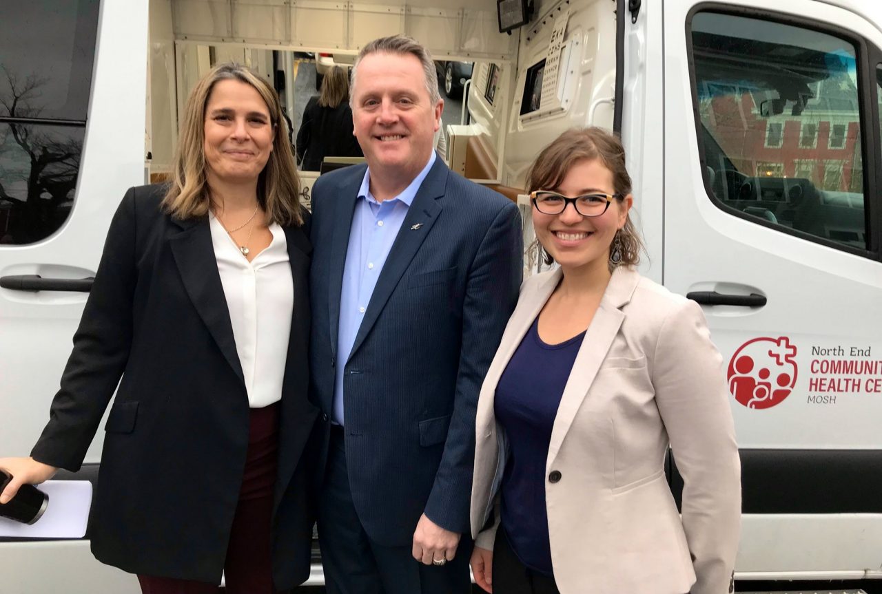 Two women and a man dressed in business casual attire smile alongside a large van with the sliding door open.