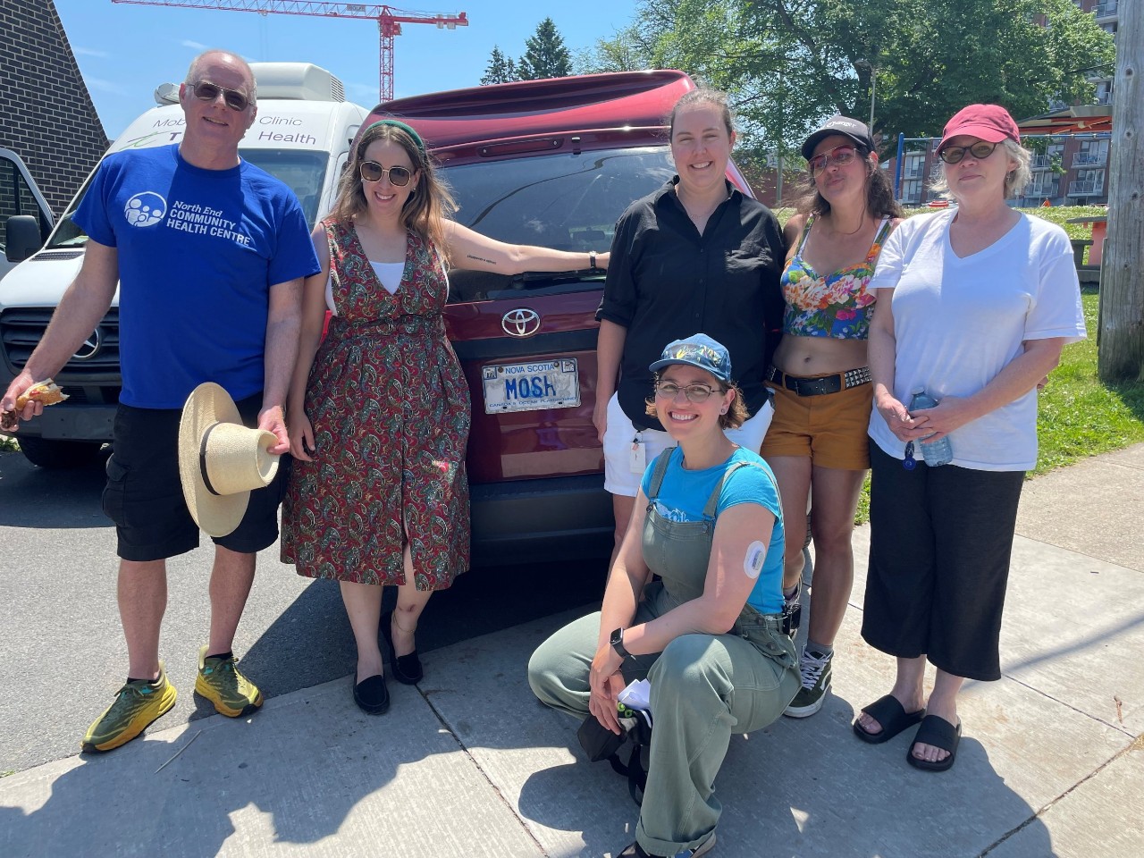 A woman in overalls, t-shirt and ball cap crouches down in front of five other people standing at the back of a red van in fine summer weather.