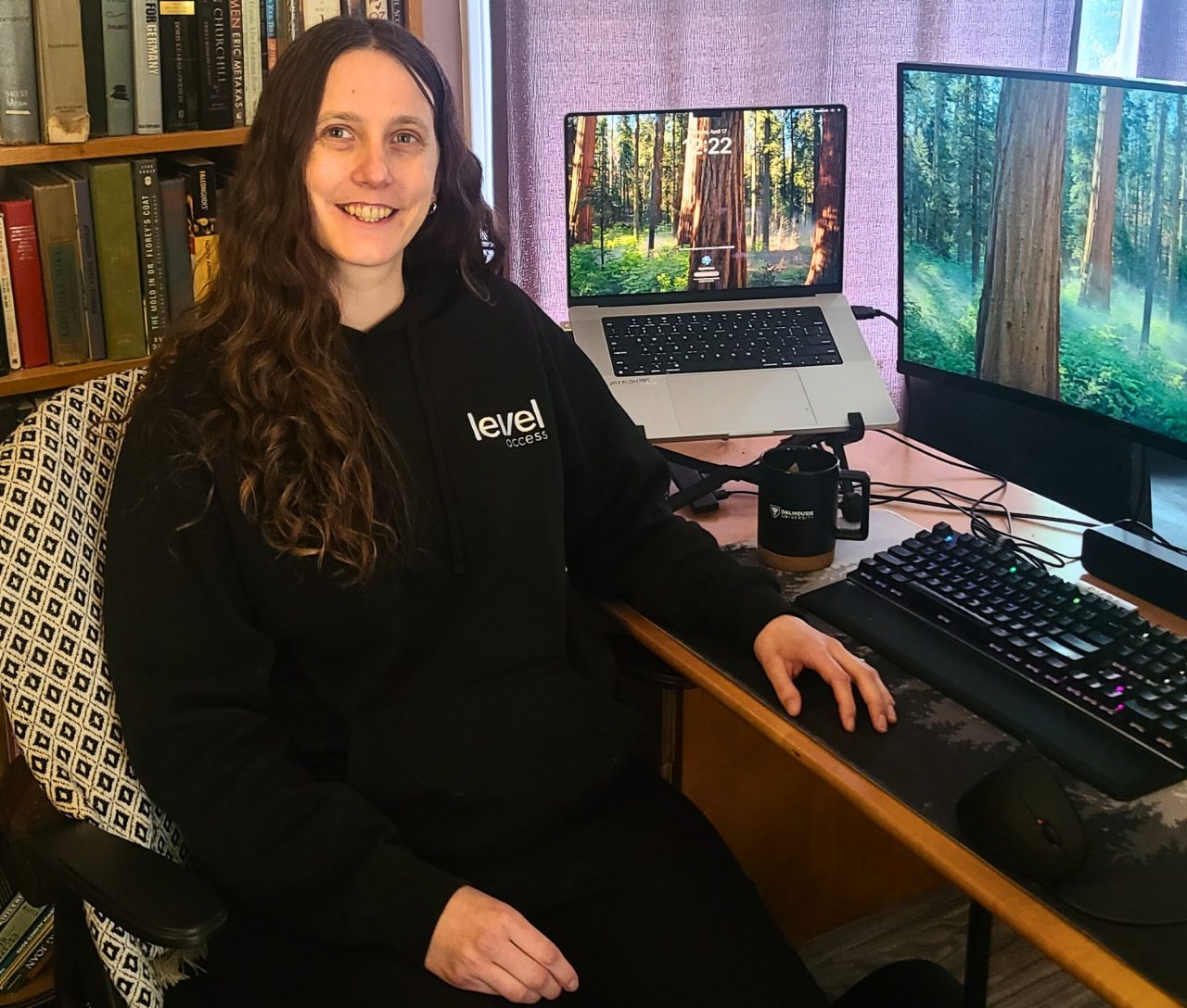 A woman wearing a black sweatshirt and pants, sitting at a desk, smiling away from her computer screens.