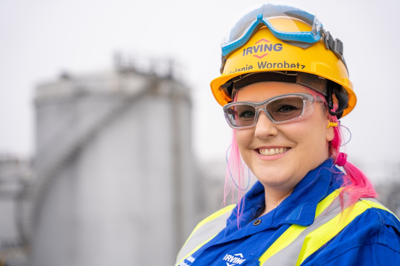 A portrait of Melanie Worobetz wearing safety glasses, a hard hat and coveralls, smiling on a work site.