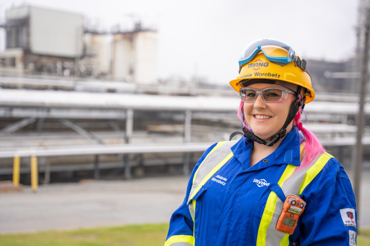 A female-presenting person with pink hair wearing safety glasses, a hard hat and coveralls, smiling on a work site.