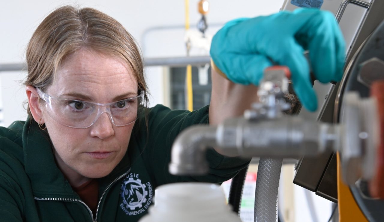 A woman wearing safety glasses and blue gloves holds a jug below a faucet about to turn on the tap as she peers intently.