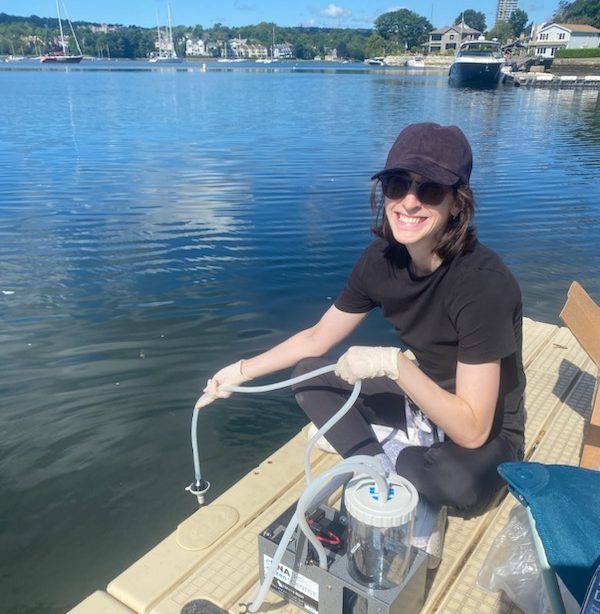 A woman wearing a ball cap and sunglasses smiles while sitting at the water's edge hanging a device into the water.