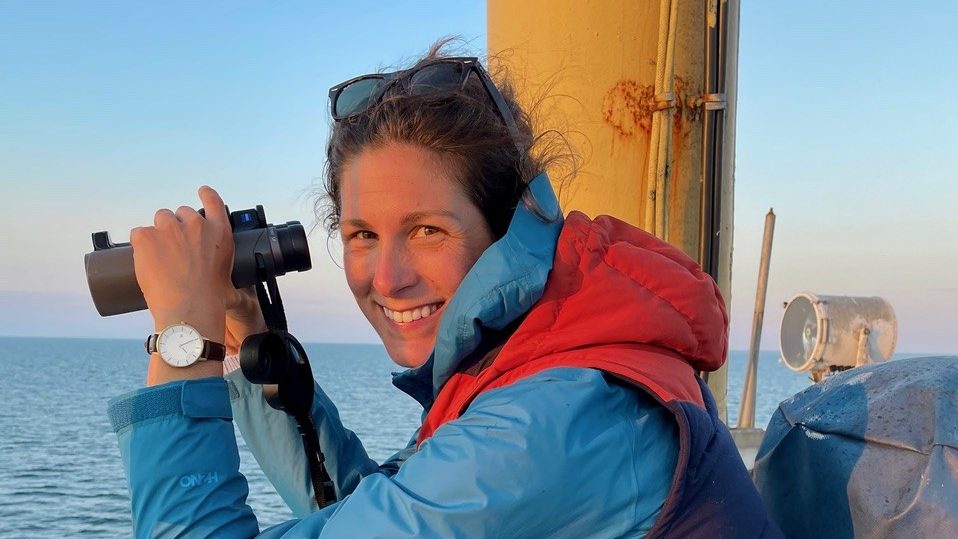 A woman smiling aboard a boat wearing a rain jacket and warm vest holding binoculars to look out to sea.