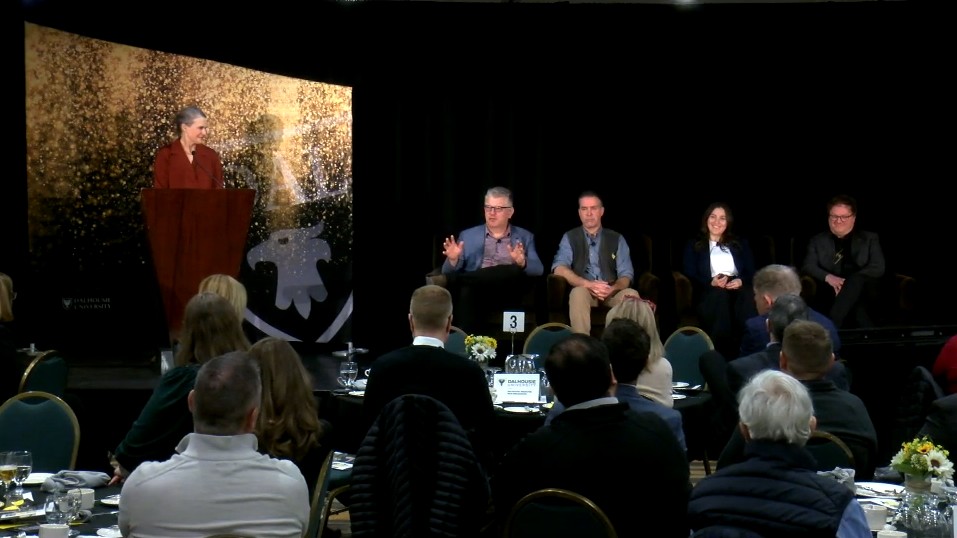 A woman standing at a podium looks to four people seated on stage next to her with an audience looking on from tables.
