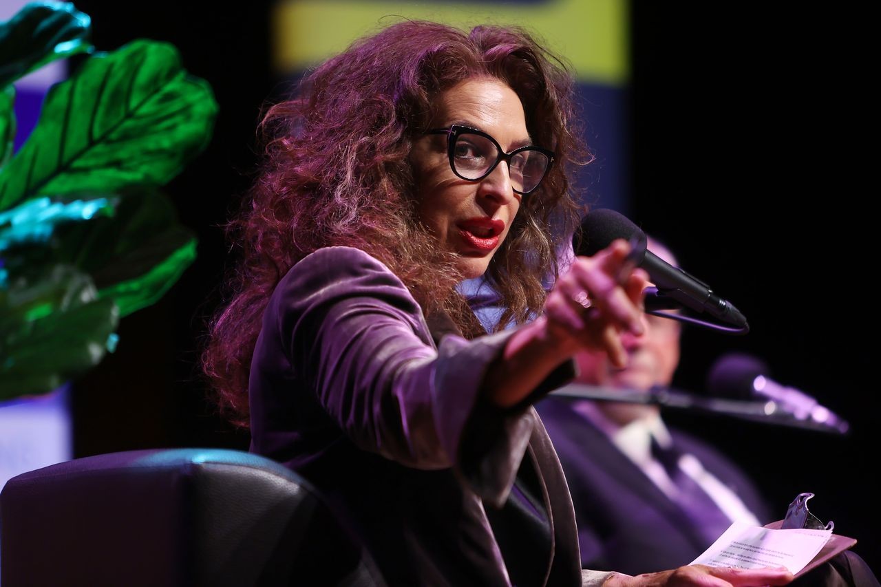 A woman points out towards the audience while seated on stage an event.