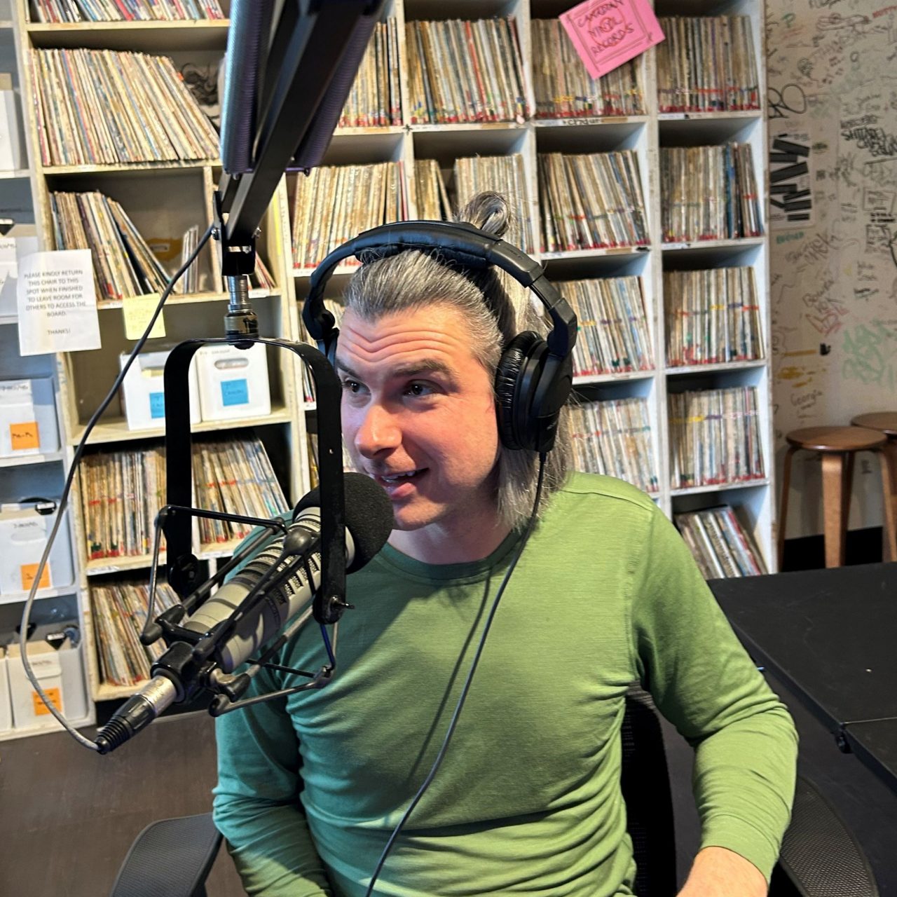 A man sits speaking into a microphone on a long arm wearing a headset in a room with shelves filled with catalogues.
