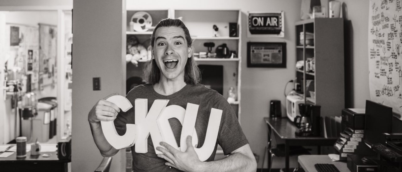 A person smiling broadly holding the letters "CKDU" in a radio studio.