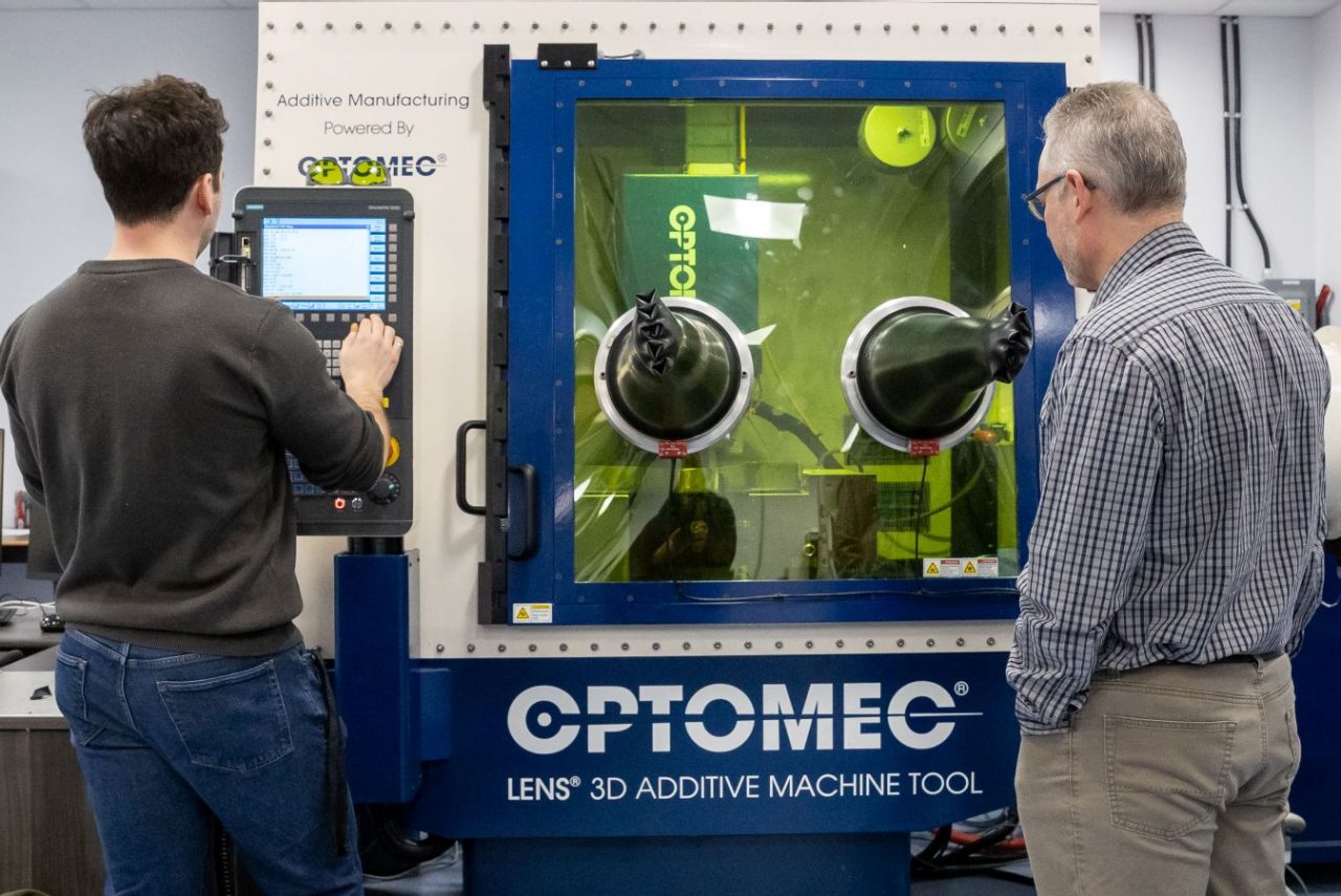 Two people stand facing a large additive machine in a lab with one working at a small screen and system.