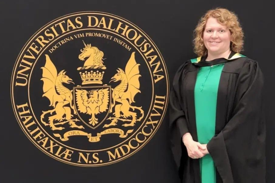 MacQueen stands with hands clasped dressed in a Convocation gown next to a backdrop with the Dalhousie University Seal. 