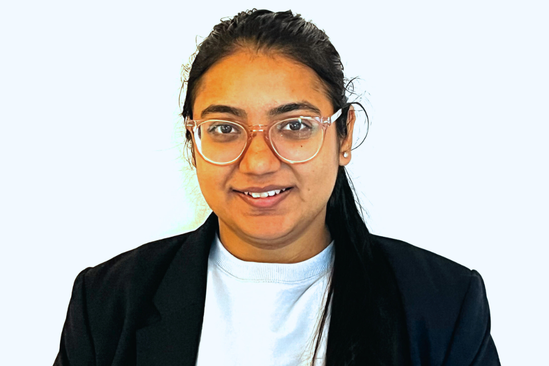 An Indian woman faces the camera straight on. Her long dark hair is pulled into a ponytail that hangs over one shoulder. She is wearing large, clear-framed glasses and a black blazer with a white T-shirt underneath. She is smiling.