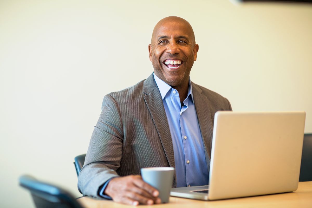 A man dressed in business attire sitting at a table with a laptop and a mug, smiling widely.
