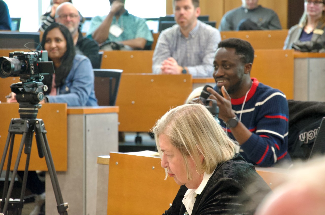 An audience member seated in a classroom at an event speaks into a microphone., smiling with other attendees in the back- and foreground.