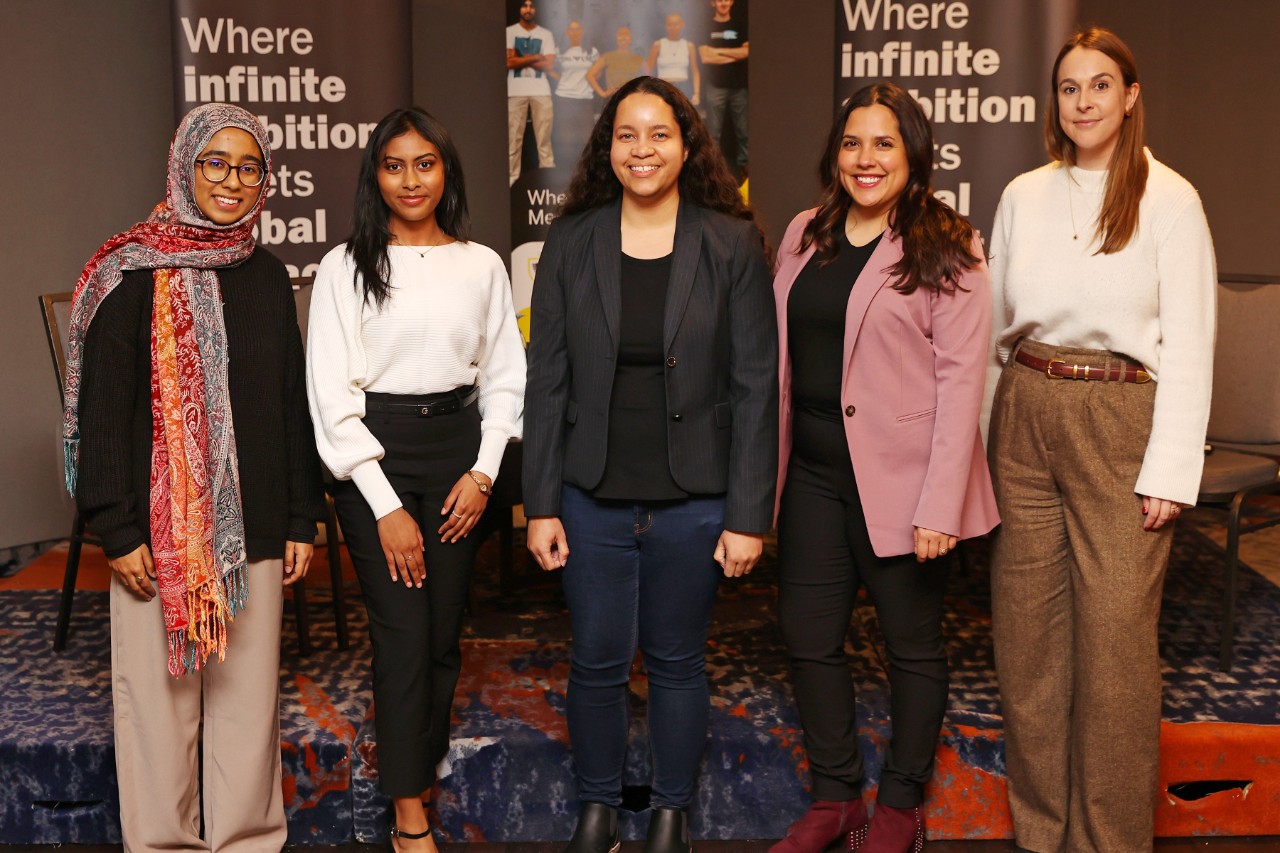 Five women of diverse backgrounds standing in a row dressed in business casual attire at an event with pop-up banners in the background.