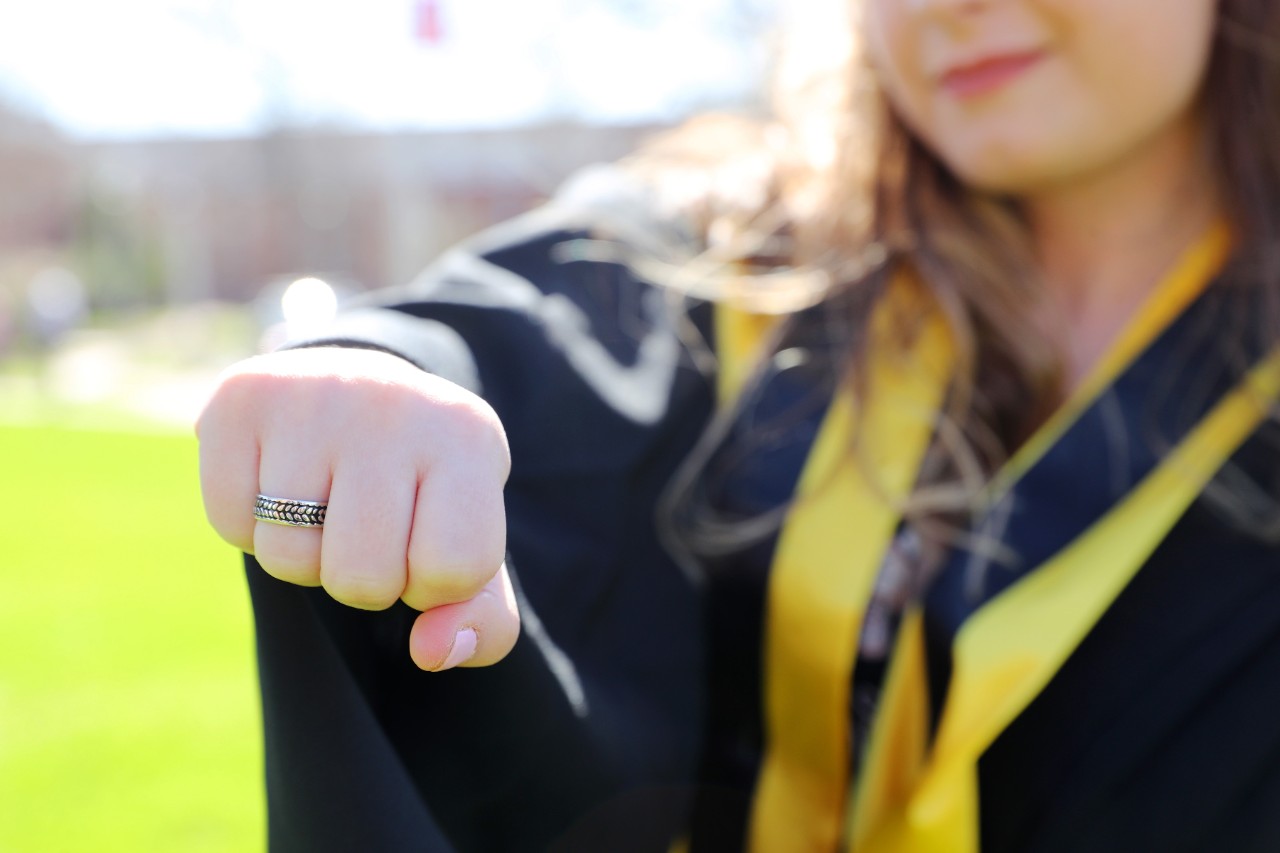 Person wearing black robes with yellow sash. Their fist is raised to show off a silver ring with a barley motif.
