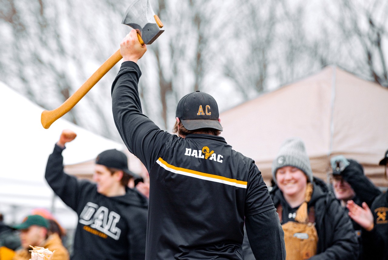 Student wearing black Dal AC jacket and hat holds axe aloft, while audience cheers in background.