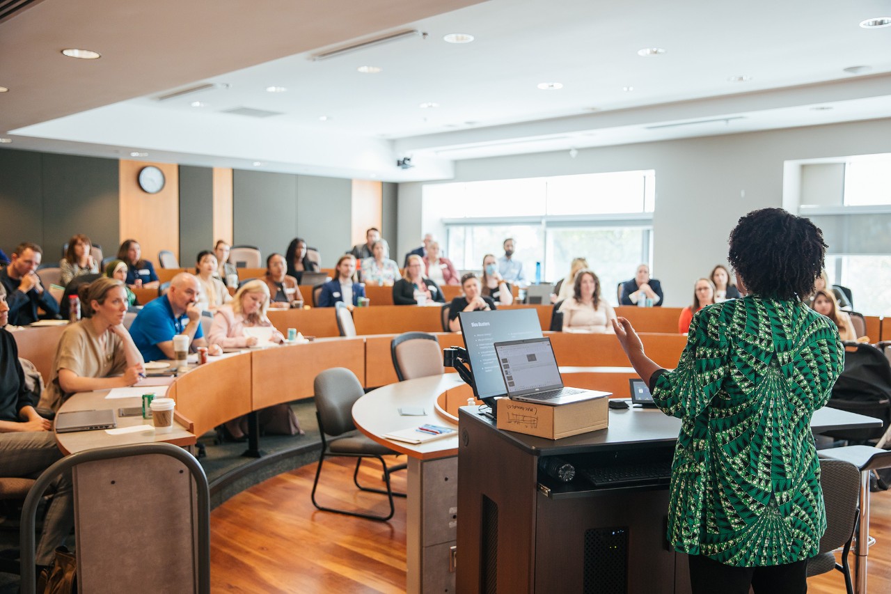 An instructor viewed from behind, with a group of students sitting at curved wood tables in a lecture hall.