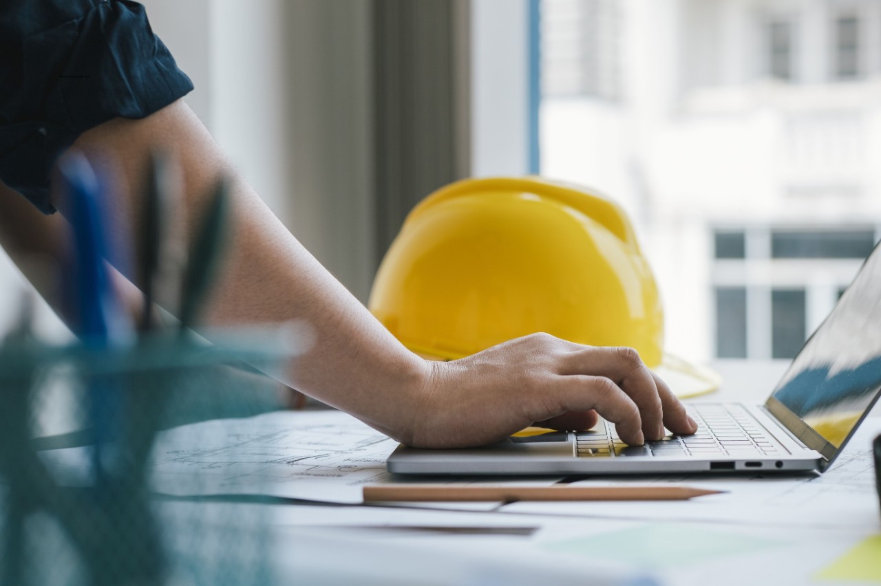 A hand types on a laptop with a yellow hard hat in the background.