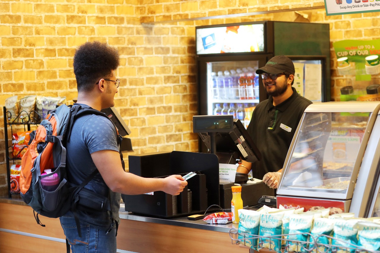 A tall male student with curly dark hair pays for a purchase at the Subway in the Killam with his DalCard