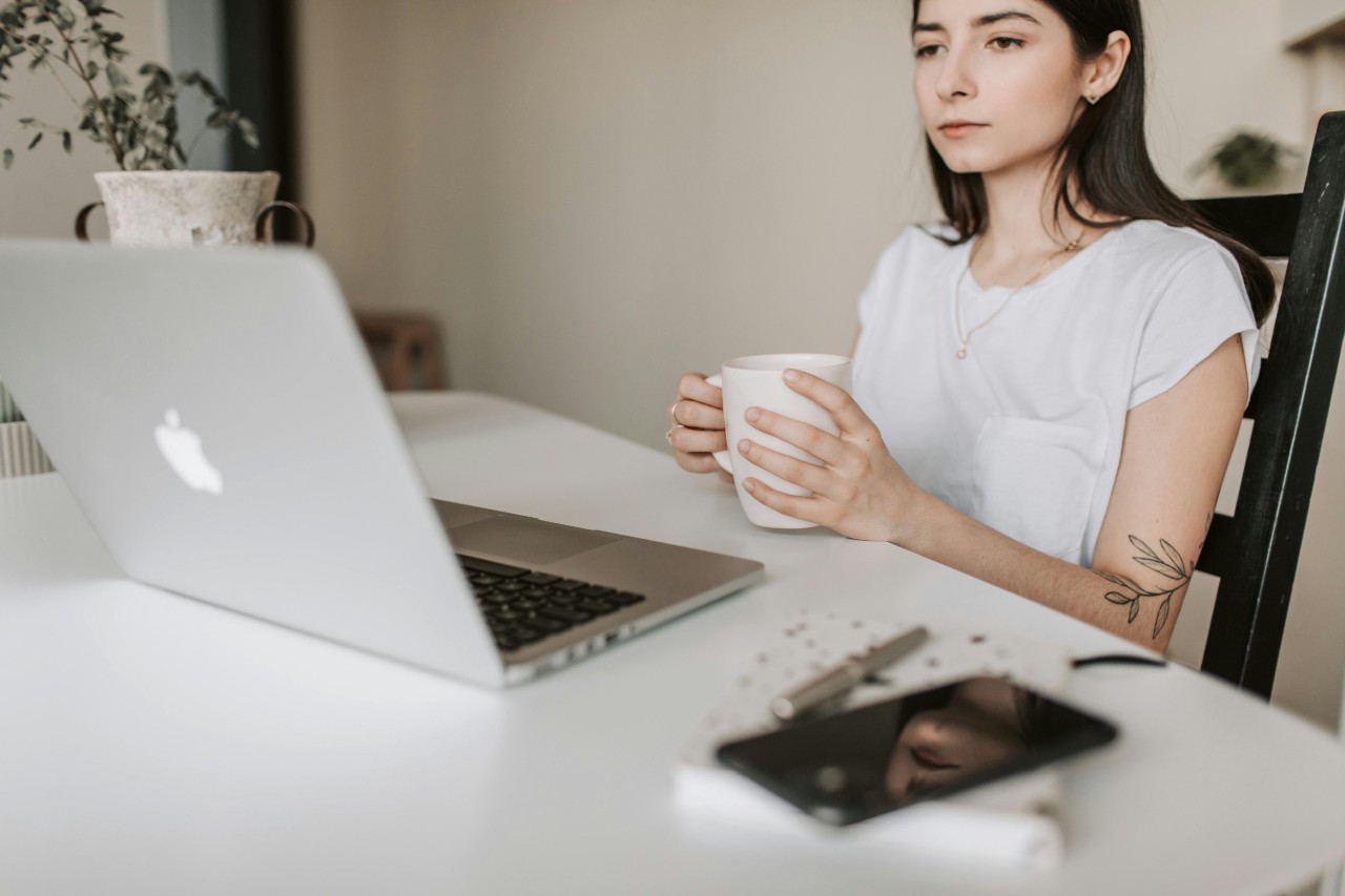 A young woman holds a coffee cup while looking at the screen on her laptop