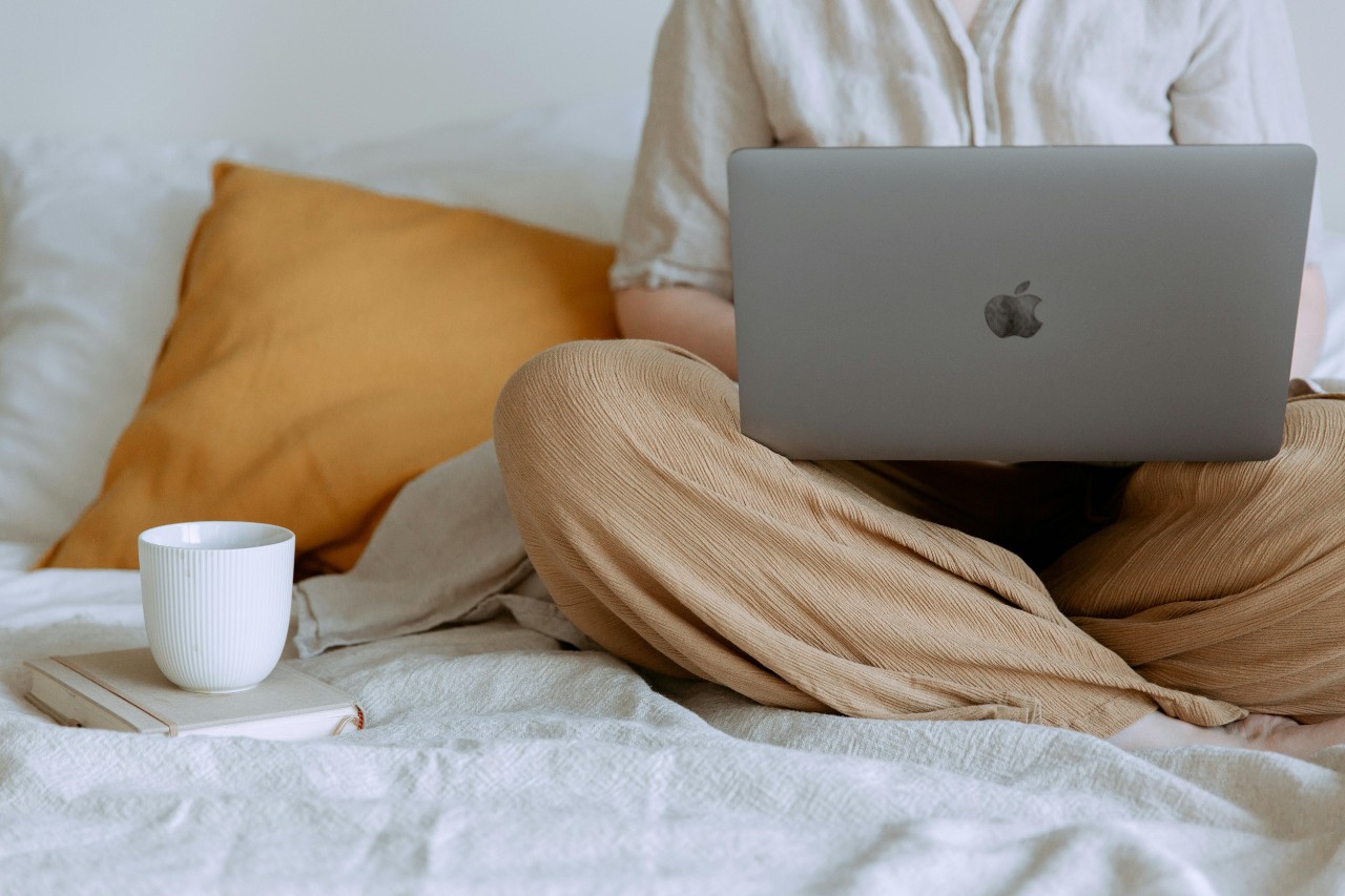 A person searches for apartment listings on a Macbook while sitting on a bed. There is a cup of coffee next to them on the bed.