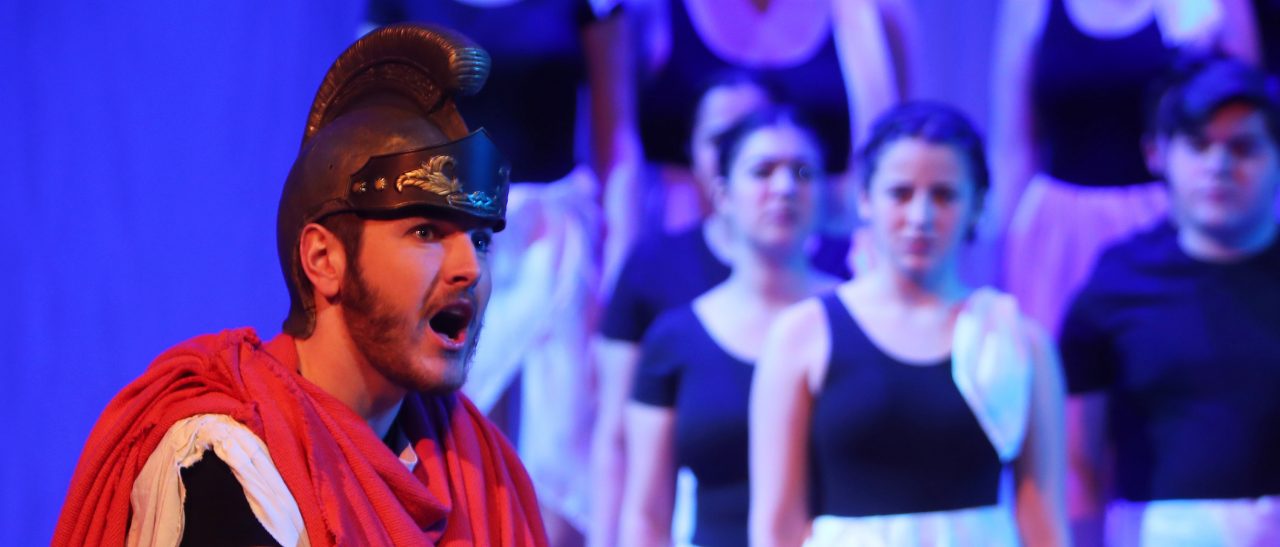 male student in costume singing at a theatre concert with dancers in the background.