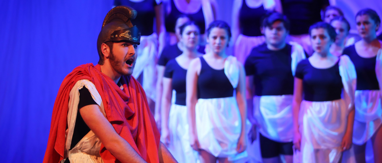 male student in costume singing at a theatre concert with dancers in the background.