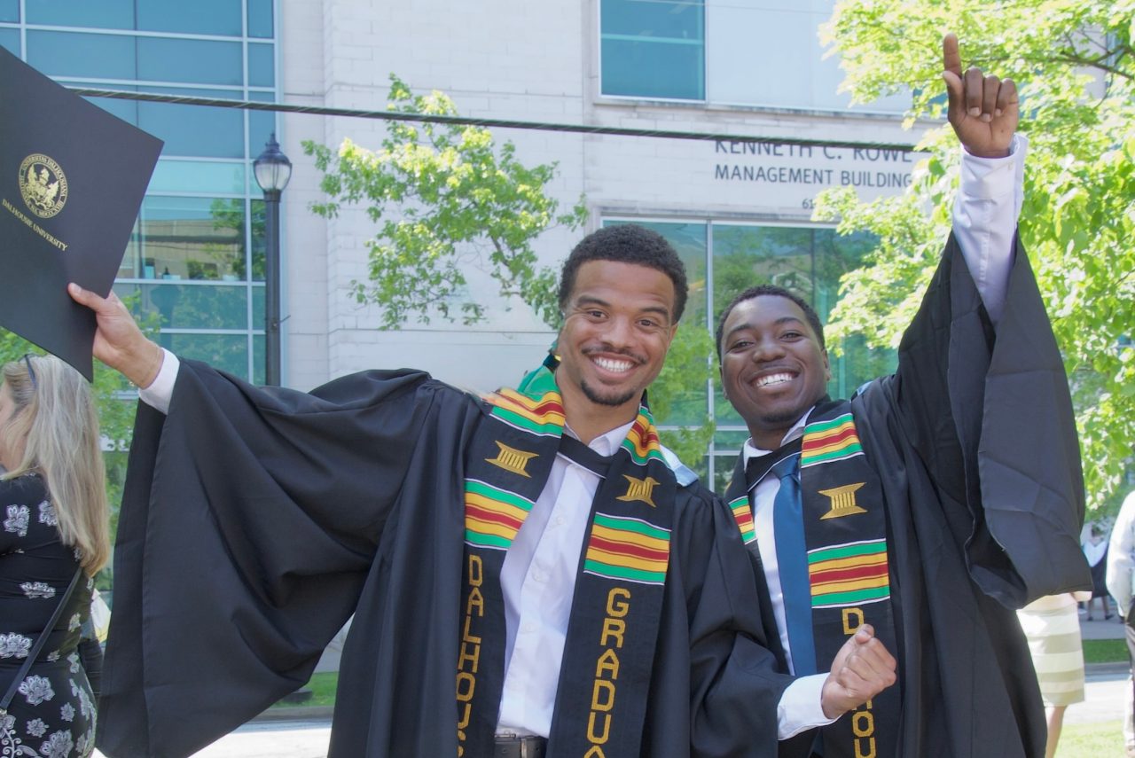 Two students wearing black robes and kente sashes raise their hands in a cheer. 