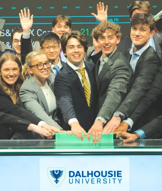 A group of students dressed in business wear cheering. They stand in front of a screen with stock market information.