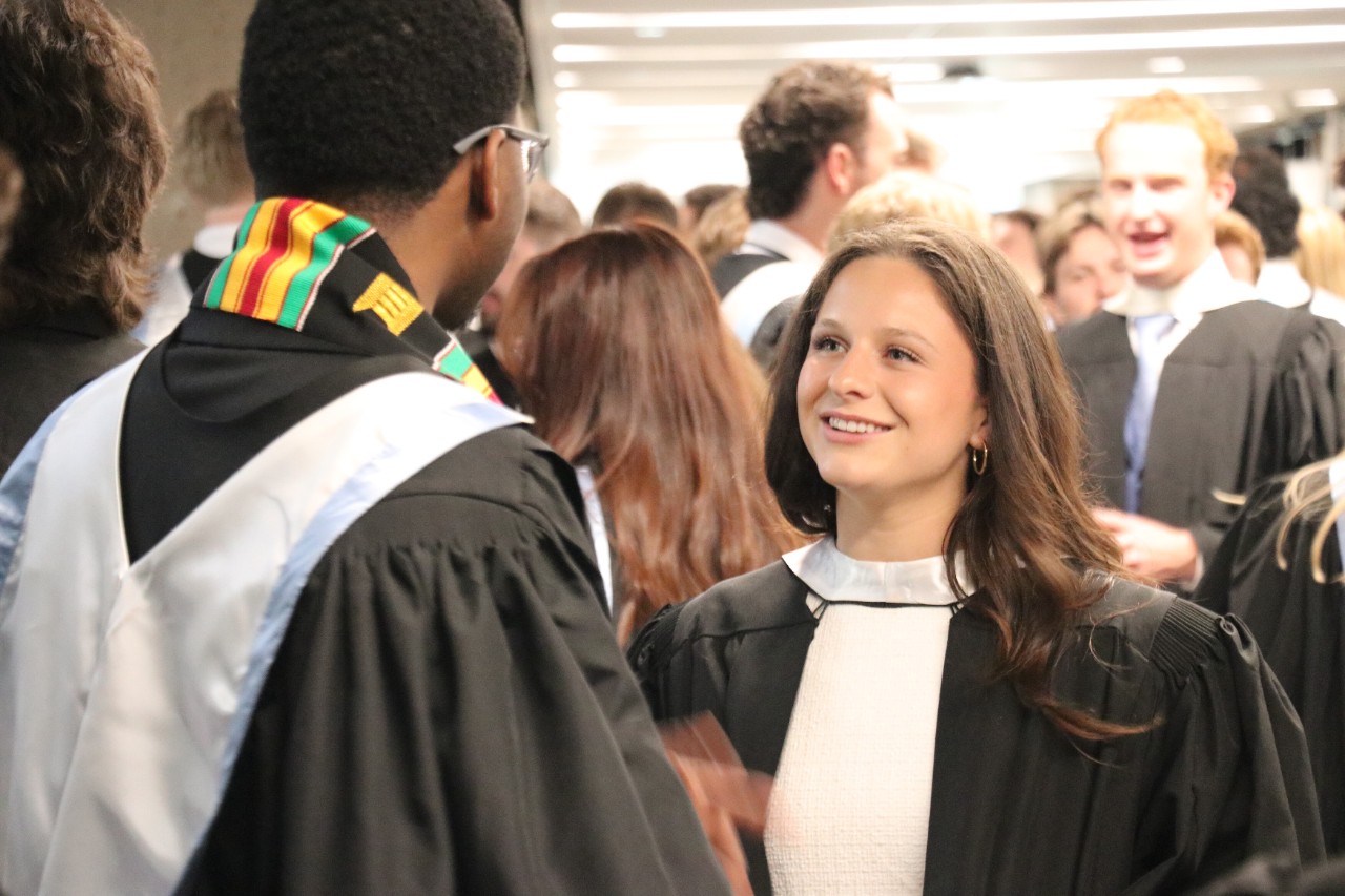 A student smiles at another student (facing away from camera). They are dressed in convcoation wear, with one wearing a kente sash in red, yellow, green and black.