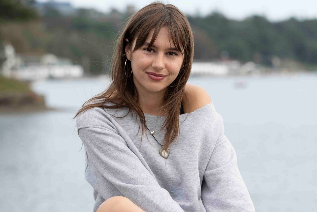 Abby sits on a rock facing the camera, smiling. In the background is water and the coastline. 