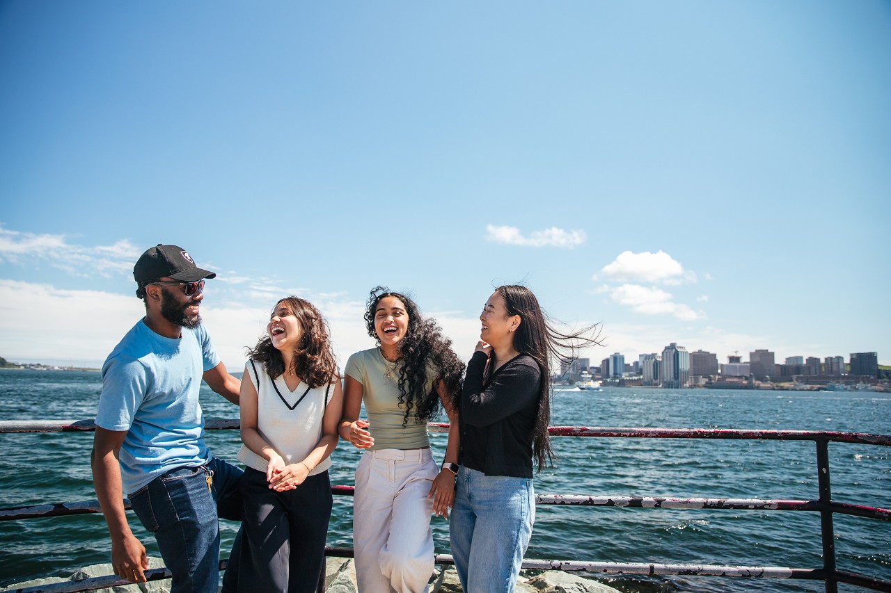 Four students laughing in the sunshine. Behind them is a body of water and a city skyline.