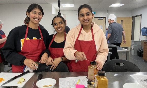 Three students wearing red aprons work together in a kitchen.