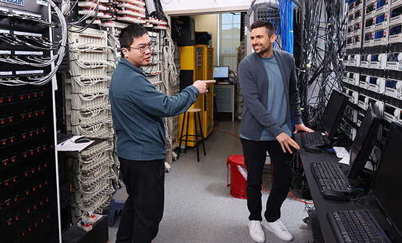 Two students work in a room surrounded by computer equipment.