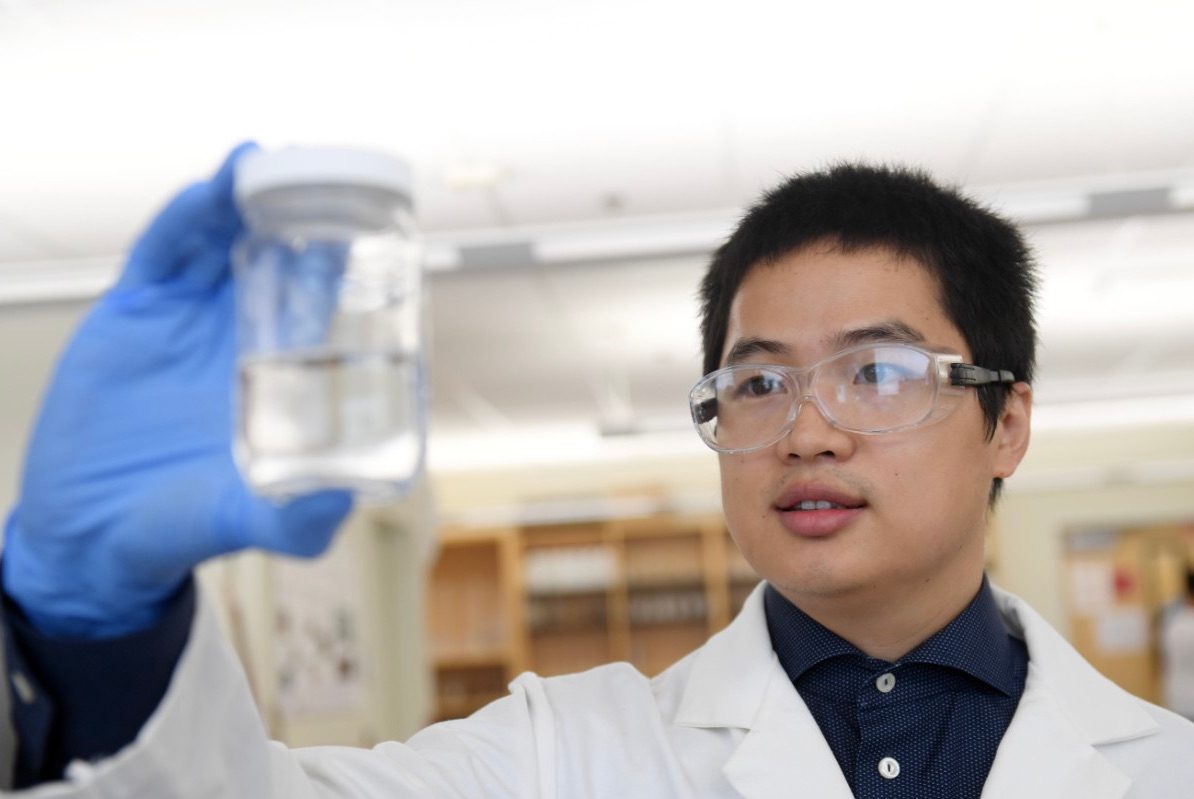 A person with short black hair is wearing laboratory goggles, a white lab coat and a blue button up long sleeve shirt.
