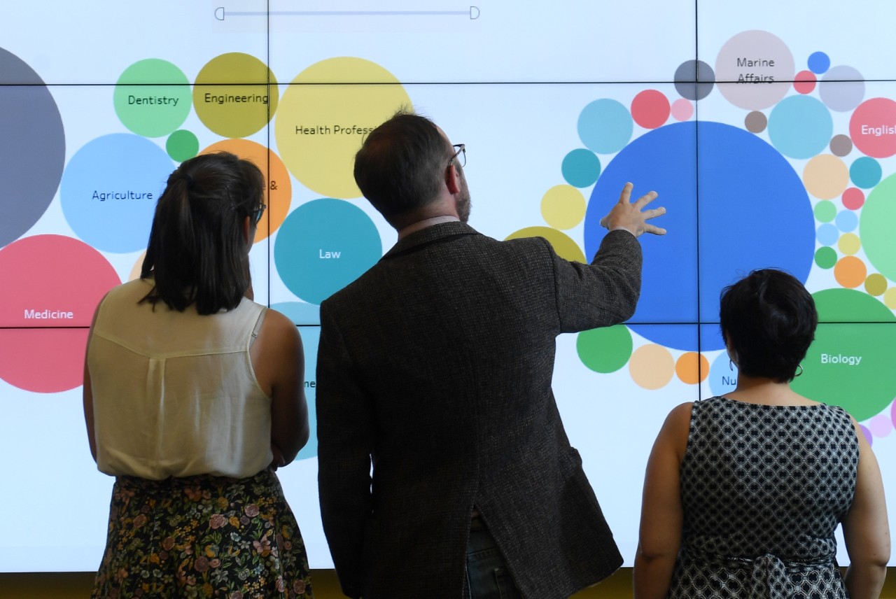 Three people stand in front of a screen with an infographic made up of several circles of different colours.