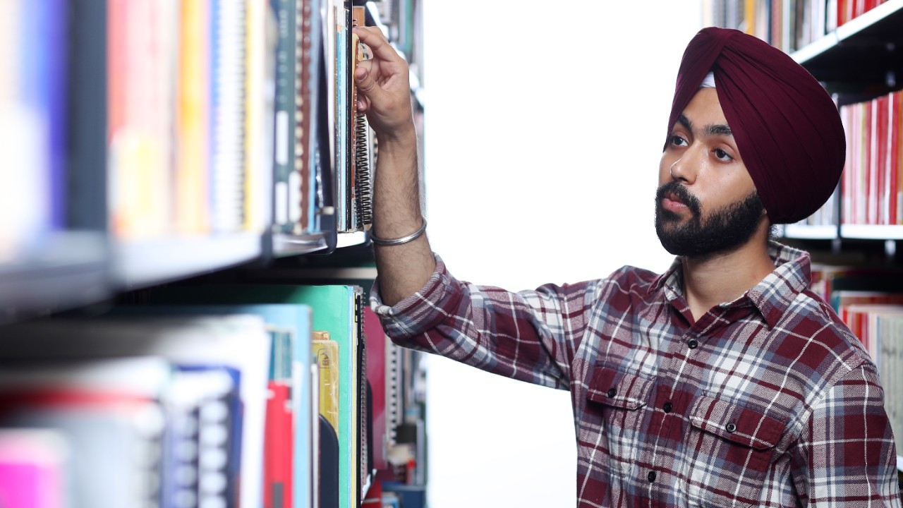 A student in a burgundy turban and shirt is reaching for a book in between two stacks of books in a library.