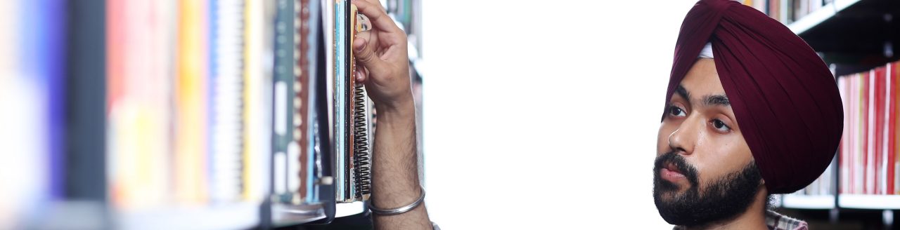 A student in a burgundy turban and shirt is reaching for a book in between two stacks of books in a library.