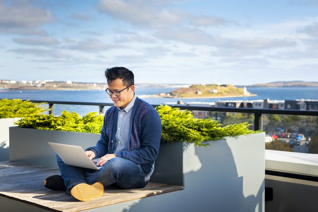 A person with short black hair sitting with a laptop with a view of the Halifax harbour.