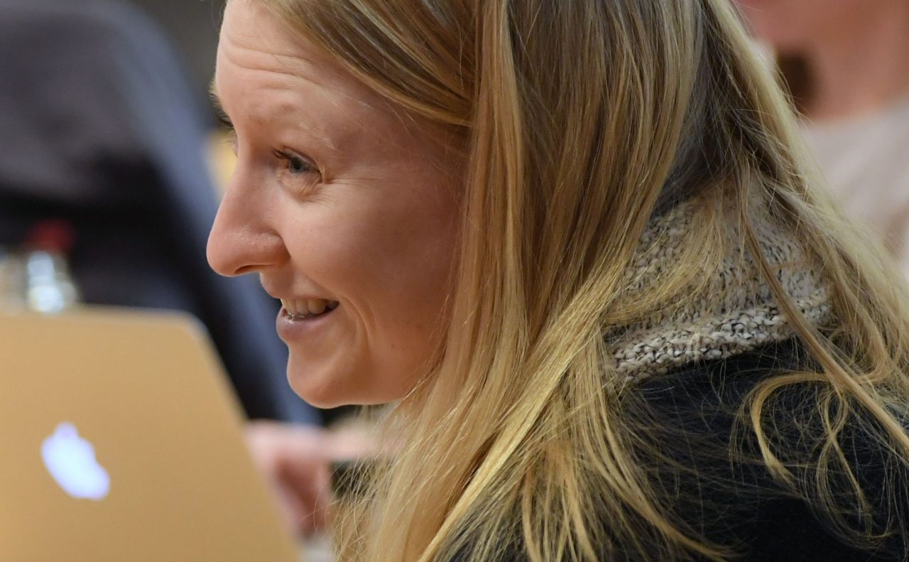 A person with long blonde hair is seen in conversation with someone out of shot.
