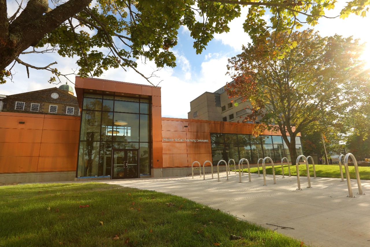An orange building, the Wallace McCain Learning Commons on a bright sunny day, with grass, trees, and bike racks in front.