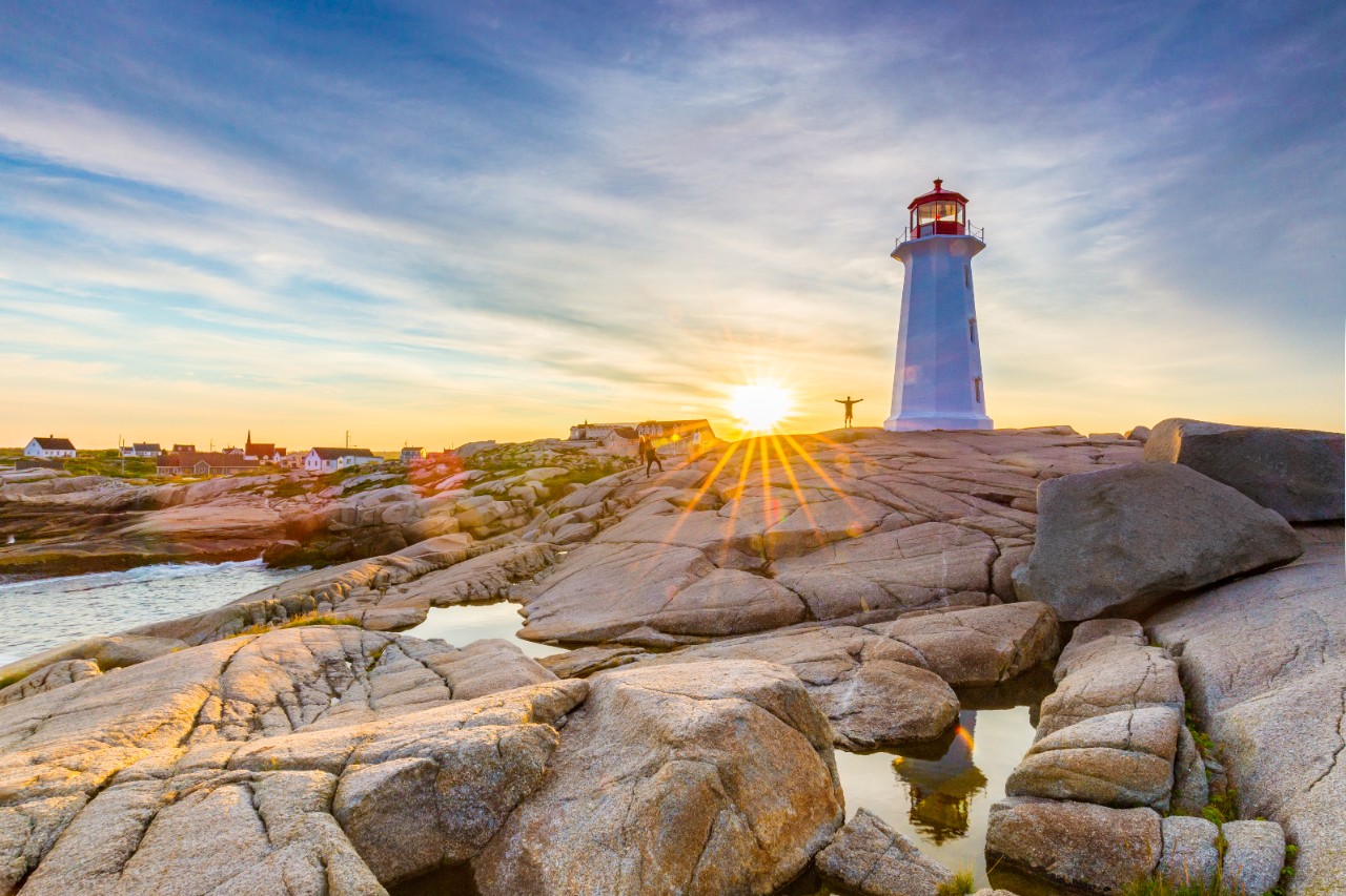Peggy's Cove, a white lighthouse with a red top is seen near sunrise from the perspective of grey rocks in the foreground.