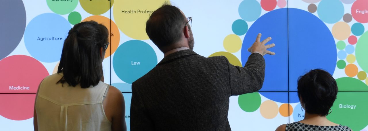 Three people stand in front of a screen with an infographic made up of several circles of different colours.
