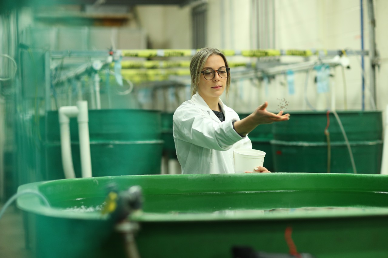 A student wearing glasses, with long blonde hair in a white labcoat in a large warehouse with large green tanks.