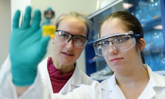 Two chemists wearing white lab coats and goggles work in a lab