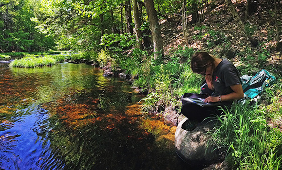 A student sits by a river, writing in a notebook