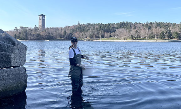 A person wearing rubber boots stands in shallow water collecting samples