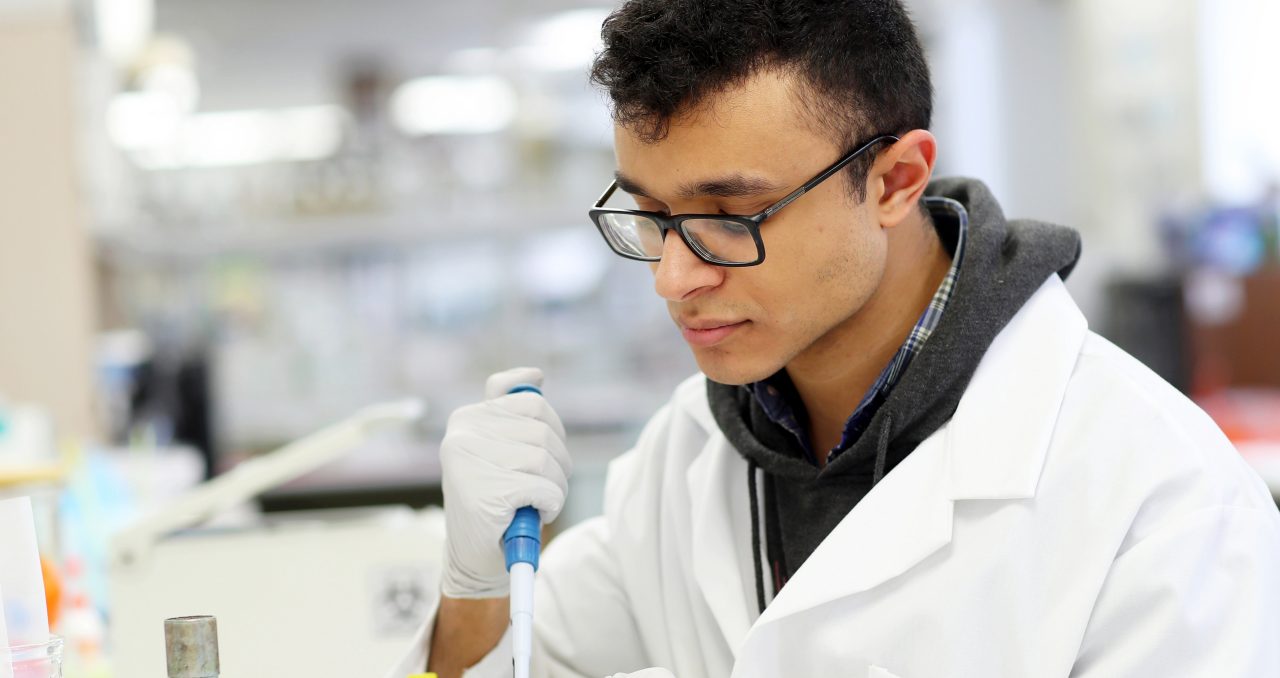 A student in a white lab coat uses a pipette to complete research