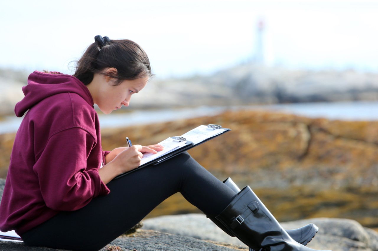 A student writes notes while they attend a field trip at Peggy's Cove, Nova Scotia. The lighthouse can be seen in the background.