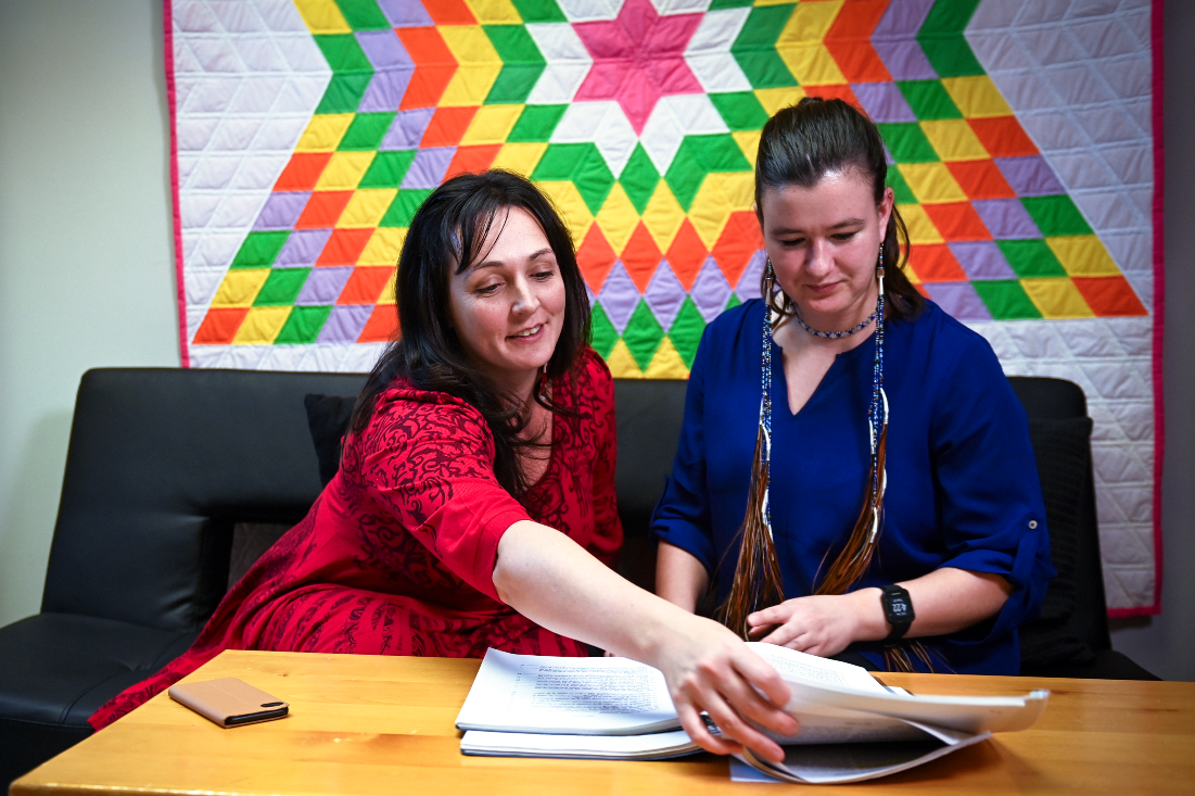 A woman wearing a red dress seated on a couch showing paperwork to a woman in a blue top 